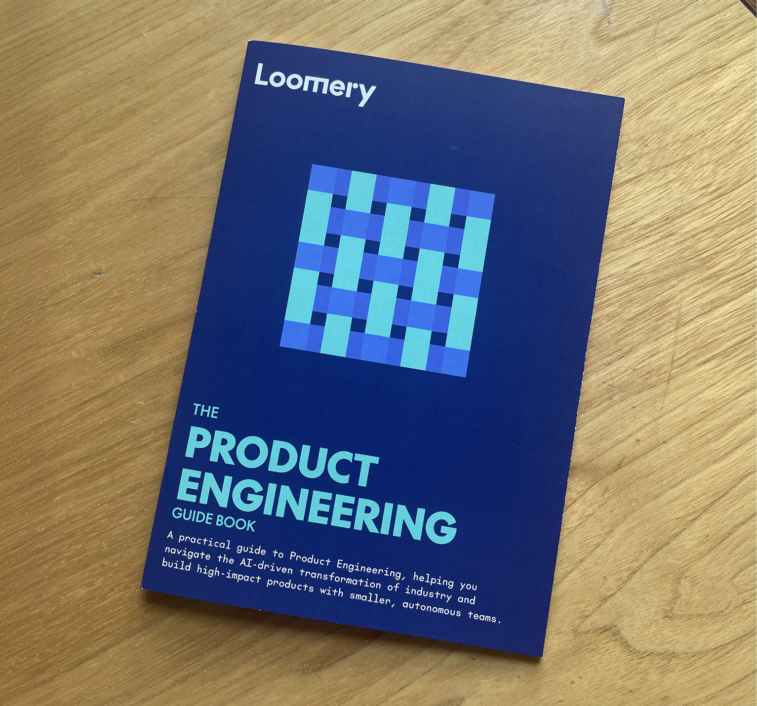 The printed Loomery Product Engineering Guidebook, with a navy cover and a teal and blue woven pattern, lying on a wooden table