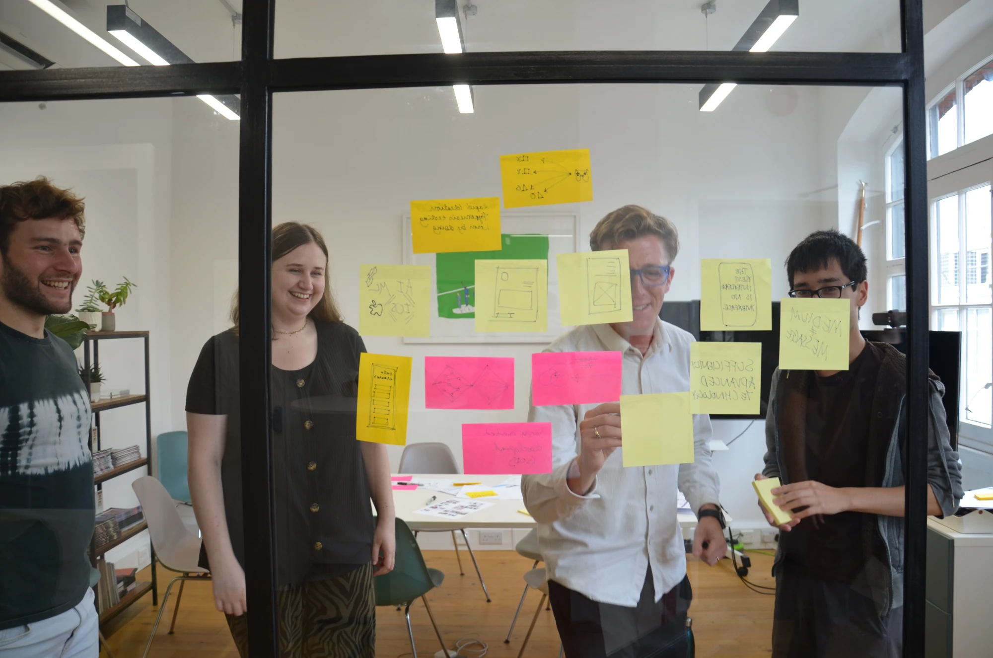 Four Loomery team members smiling as they sketch ideas on sticky notes stuck to a glass wall