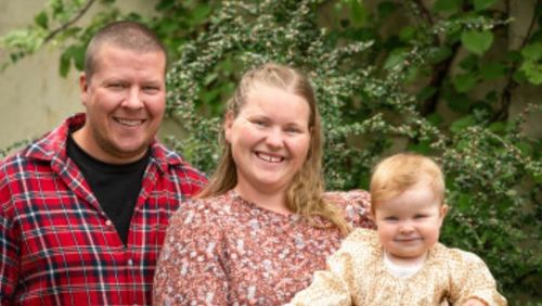 Foto: Kommuneverter Anna og Jørgen Bergo står foran en vegg av grønne planter og smiler til kamera med et lite barn i armene.