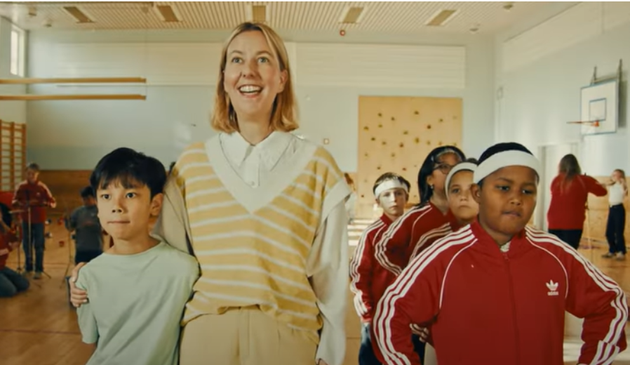 Stilbilde om foreldrefrivillighet. Mor står med sønn i gymsal full fra barn fra samme lag .