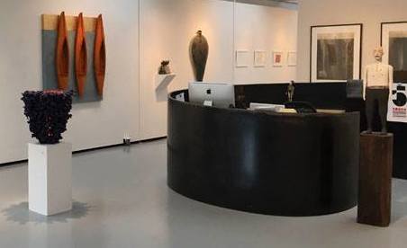 Art gallery interior featuring a black curved reception desk surrounded by various artworks. Image courtesy of Joy Tahan Ruddell.