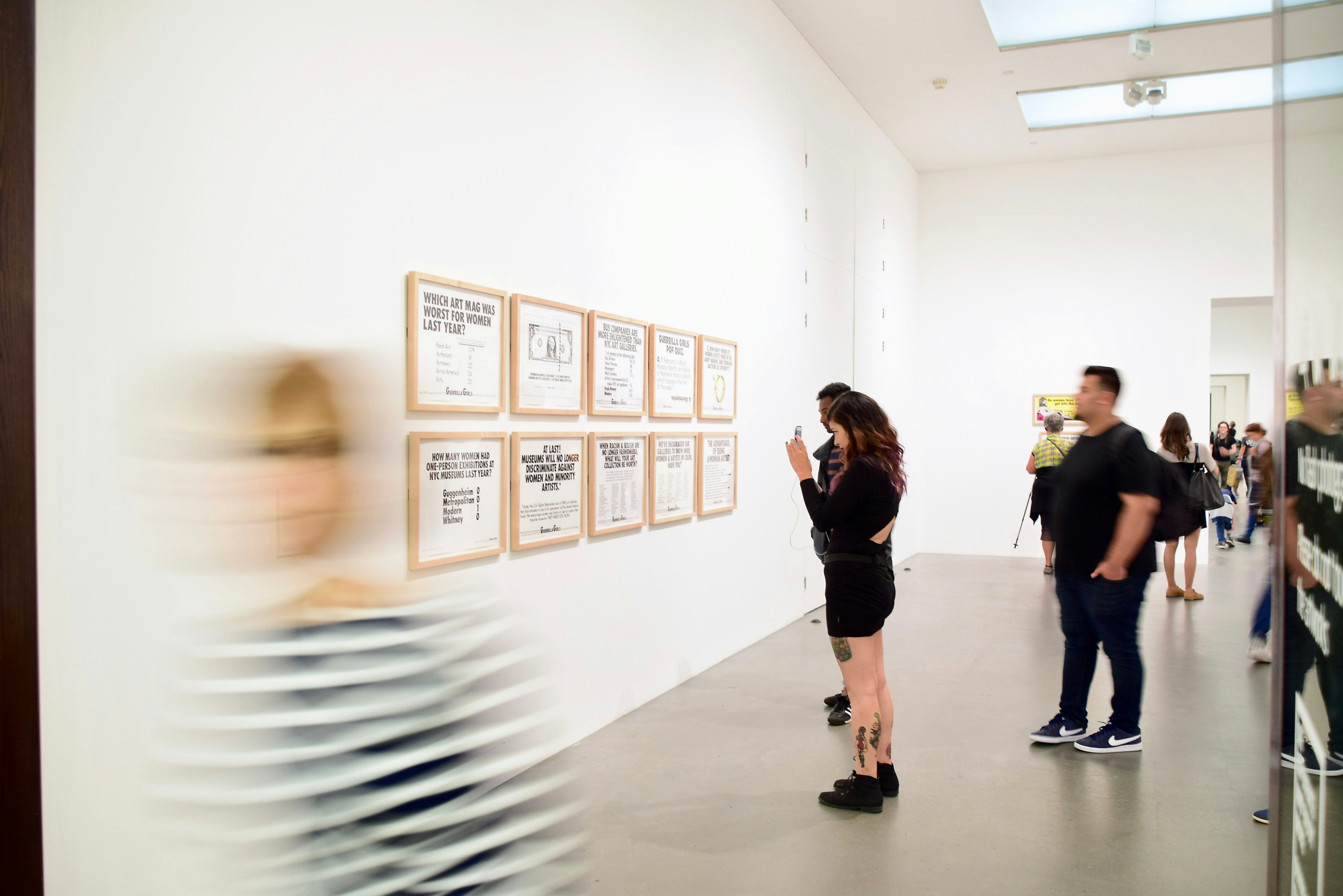 A blurred figure walks past framed text-based artworks on a white gallery wall. Image courtesy of iSAW Company.