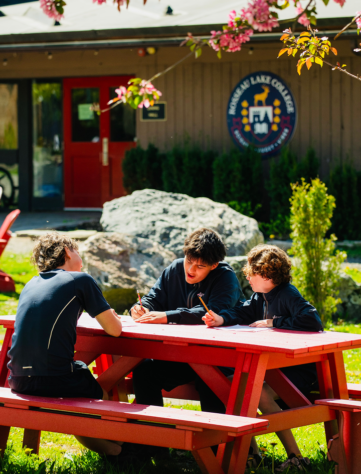 3 teenage boys sitting on a table with Rosseau Lake College logo on the back of a shack