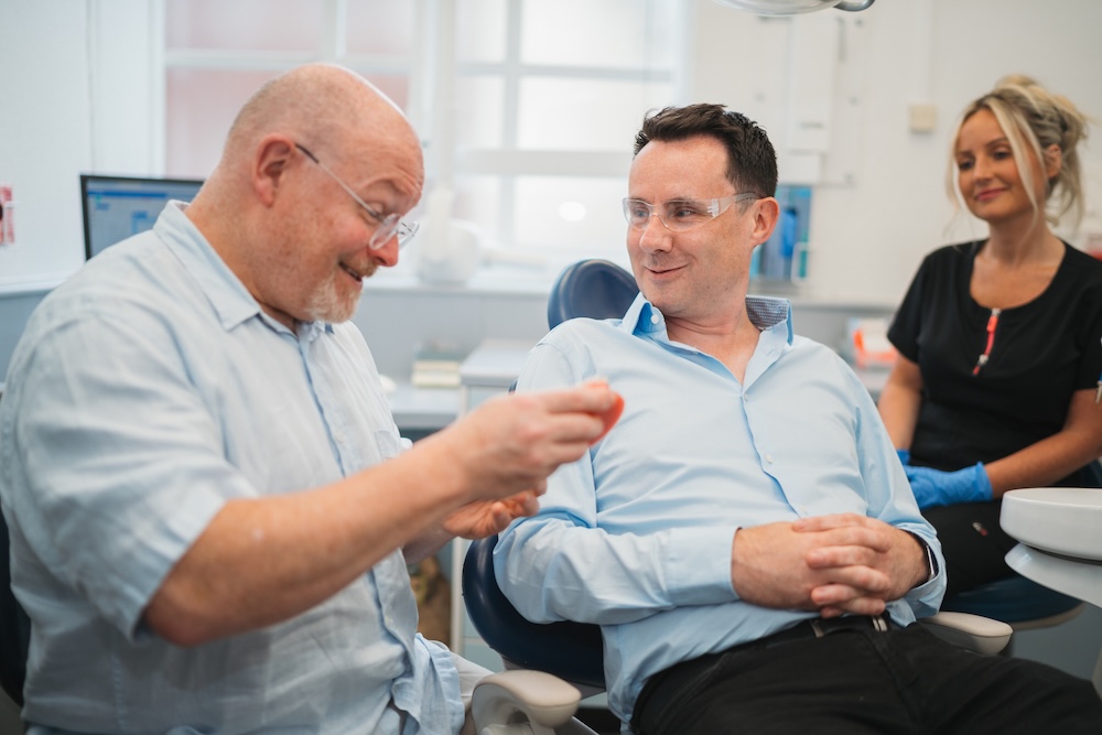 A dentist explaining a dental model to a male patient wearing protective glasses, with a female dental assistant watching.