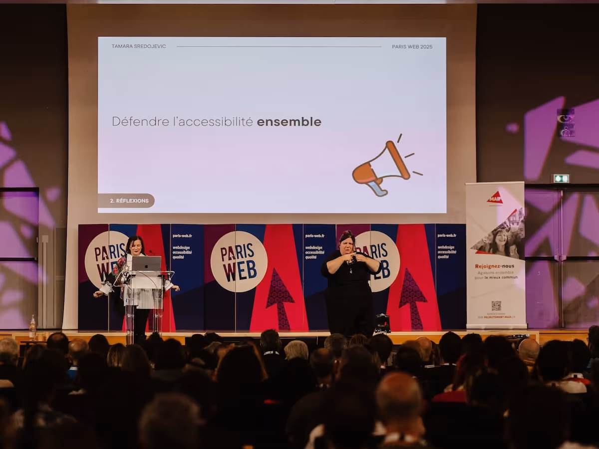 Tamara standing on Paris Web stage in front of a slide that reads "Defending accessibility together"