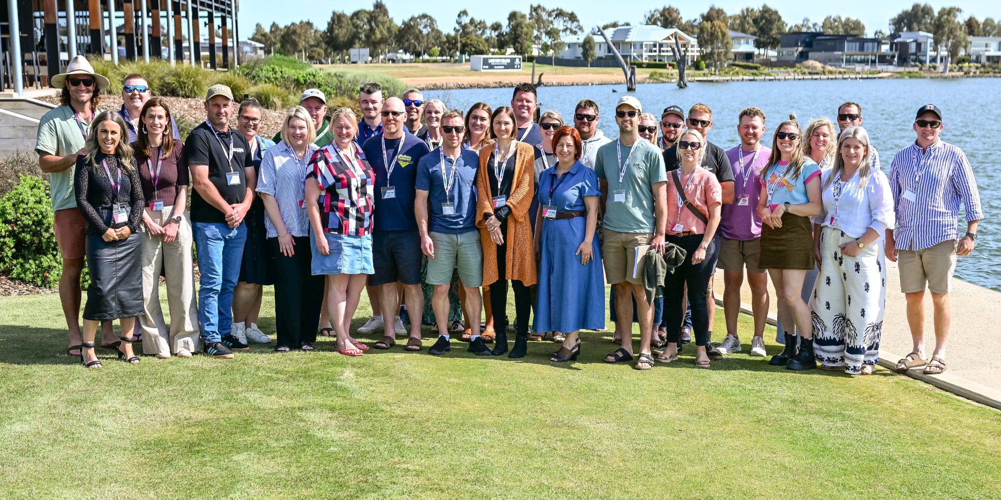 A large group of tradie business owners and their partners smiling together outdoors at a Ladies with Tradies event, gathered on a waterfront lawn on a sunny day.
