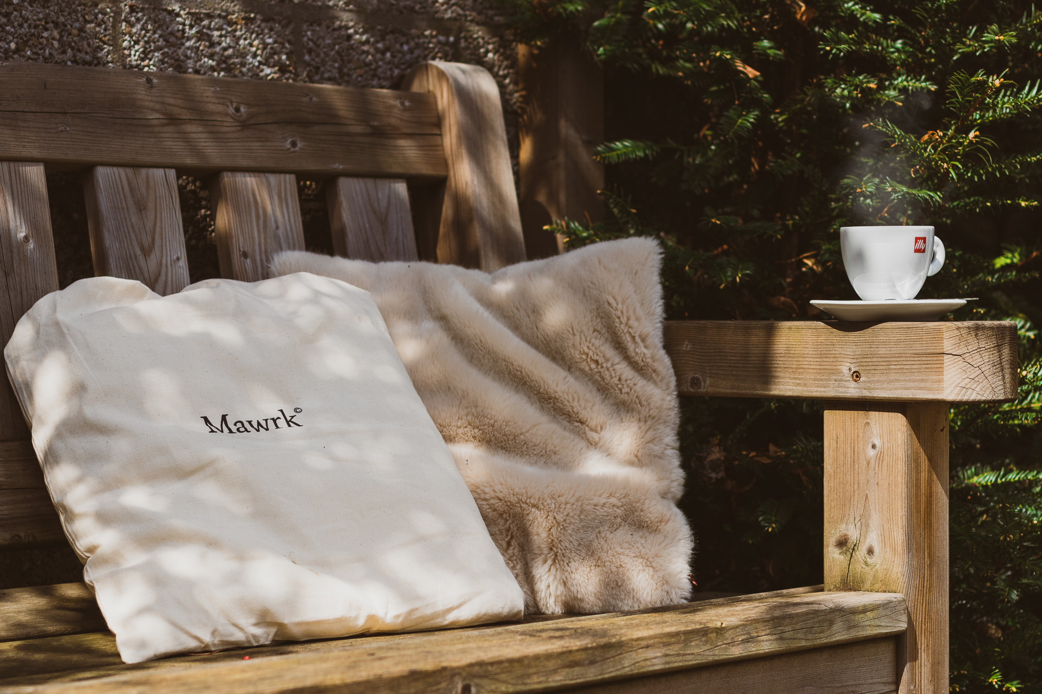 A cotton pouch with Mawrk logo on it, used as pillow, together with another fluffy pillow on a wooden bench accompanied by a steaming cappuccino cup.