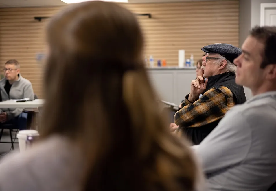 Group of adults sitting in a classroom or meeting room, attentively listening, with focus on an older man wearing a flat cap and plaid shirt.