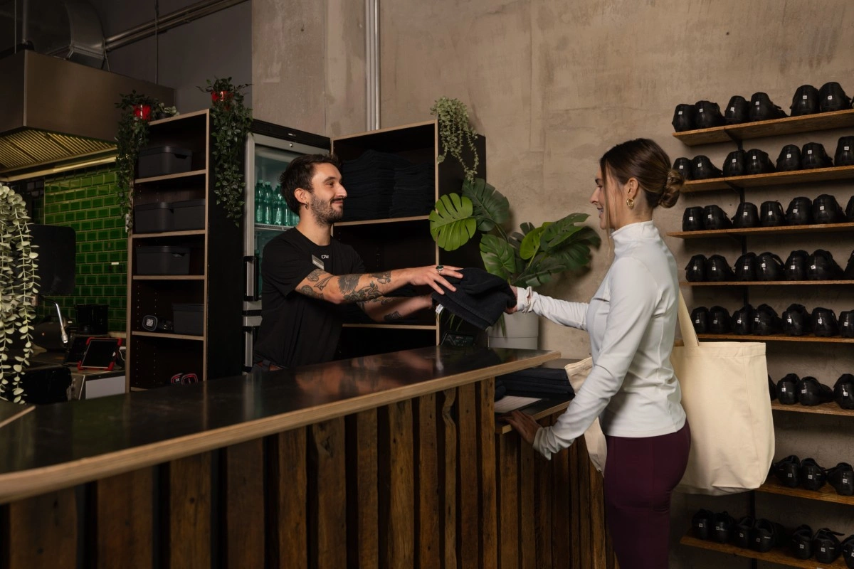 Man with tattoos handing folded black clothing to a woman with a white shirt and large tote bag across a wooden counter in a modern retail store.