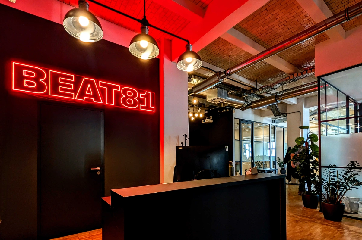 Modern office reception area with a black desk, glass partitions, indoor plants, and a red neon sign reading 'BEAT81' on a dark wall.