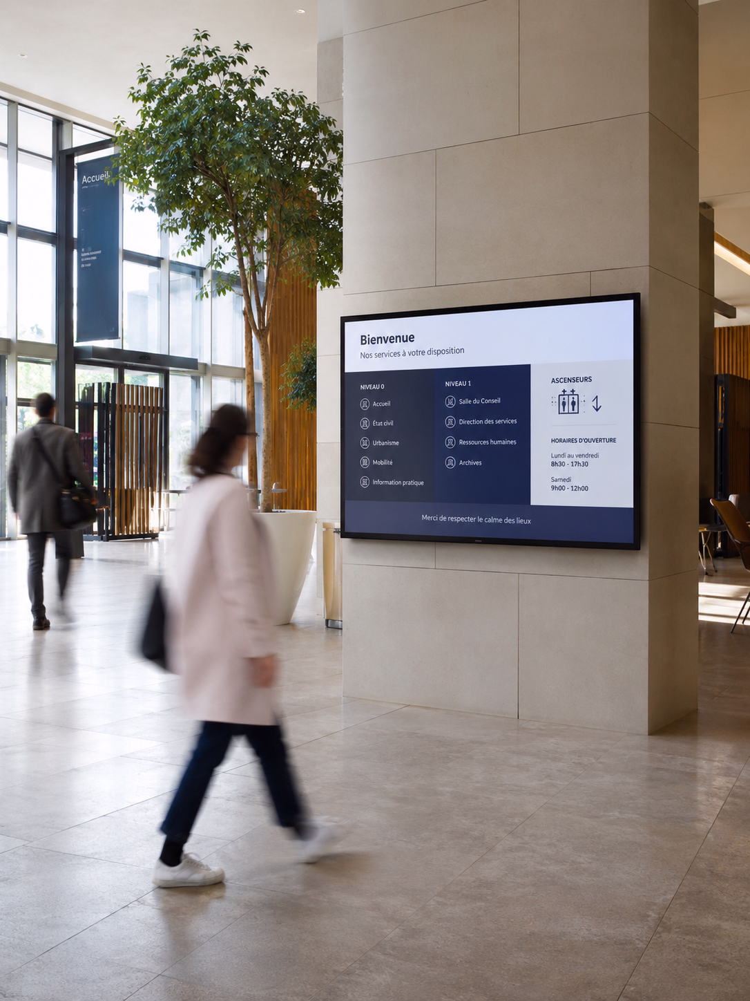 Écran d’affichage dynamique 16:9 dans le hall lumineux d’une métropole française, diffusant une signalétique vers les différents services du bâtiment, avec des usagers en mouvement.