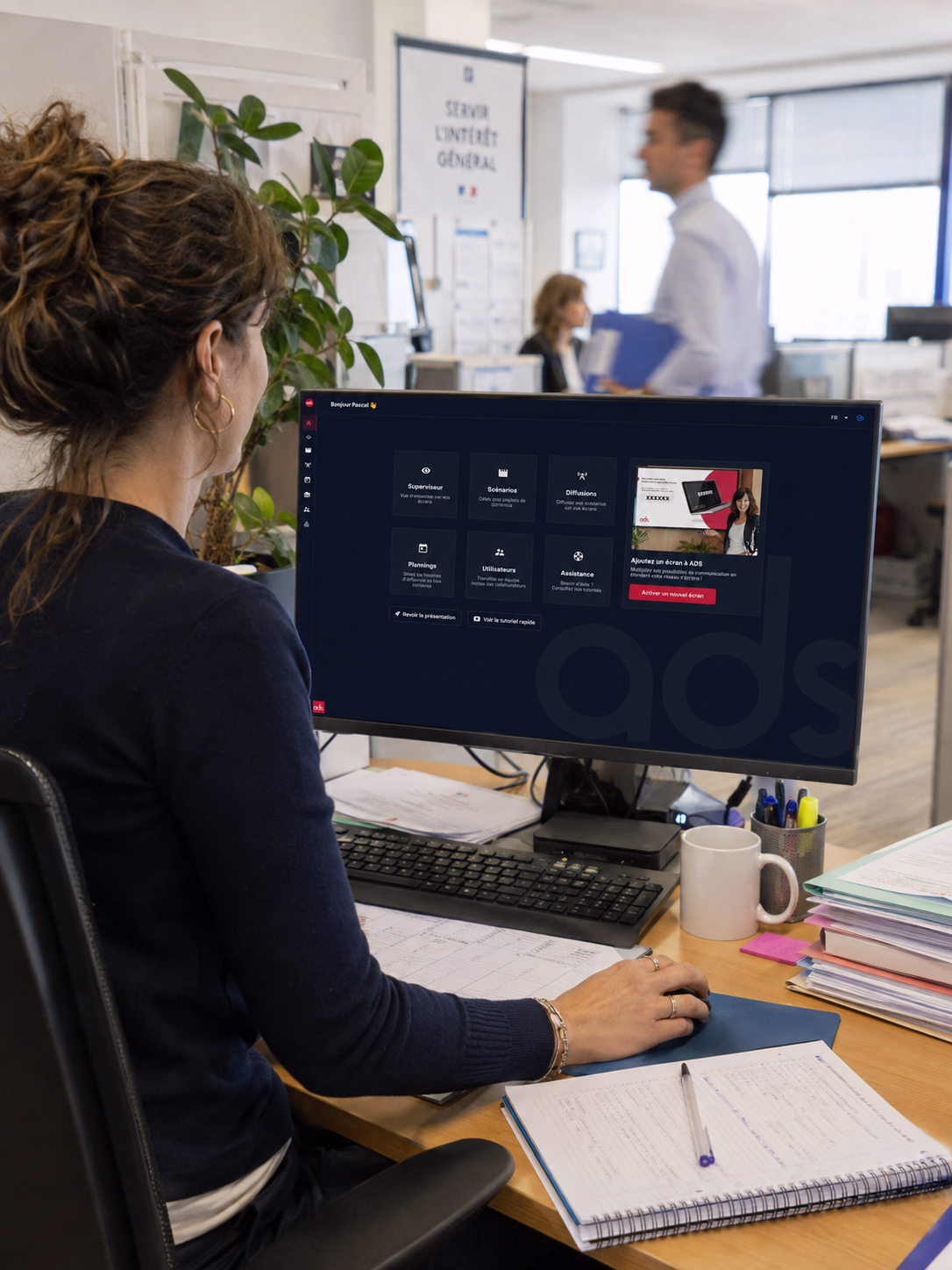 Fonctionnaire française travaillant sur un ordinateur dans un bureau administratif lumineux, avec l’interface ADS affichée à l’écran et des collègues en mouvement en arrière-plan.