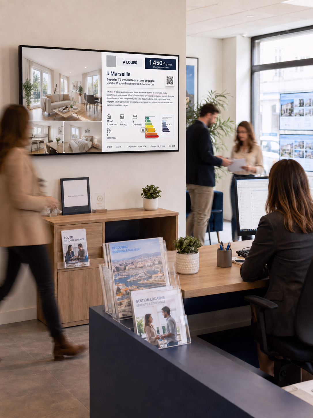Écran d’affichage dynamique installé dans les bureaux d’une société de gestion locative, diffusant une annonce de location pour un appartement à Marseille avec photos intérieures, loyer, caractéristiques du bien, QR code et diagnostics énergétiques, tandis que des collaborateurs travaillent autour et que certaines personnes apparaissent en mouvement flou.