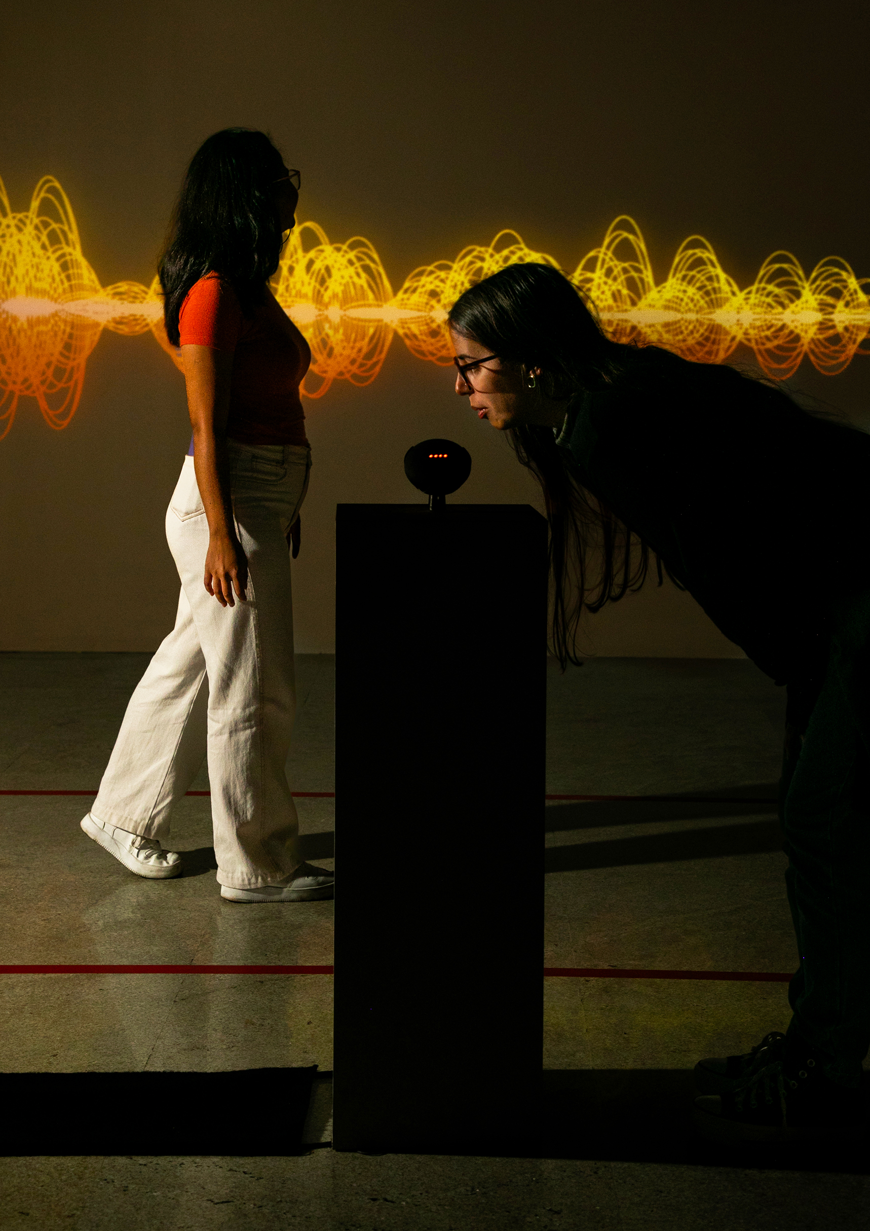 Two visitors interacting with a small device on a pedestal while a yellow waveform-like projection stretches across the wall behind them.