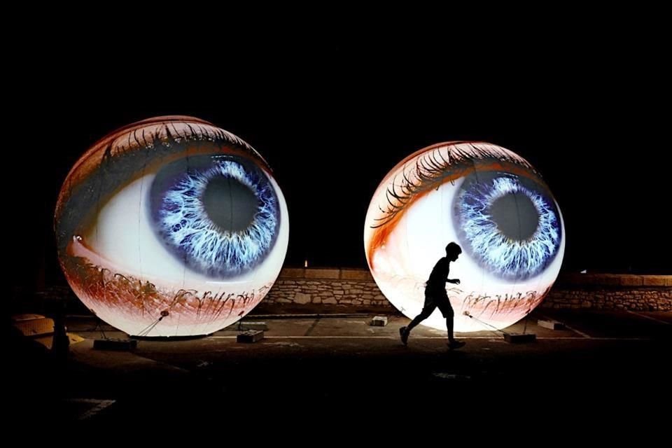 Two enormous illuminated sculptures of blue human eyes against a black night sky, with a person silhouetted running in front of them.