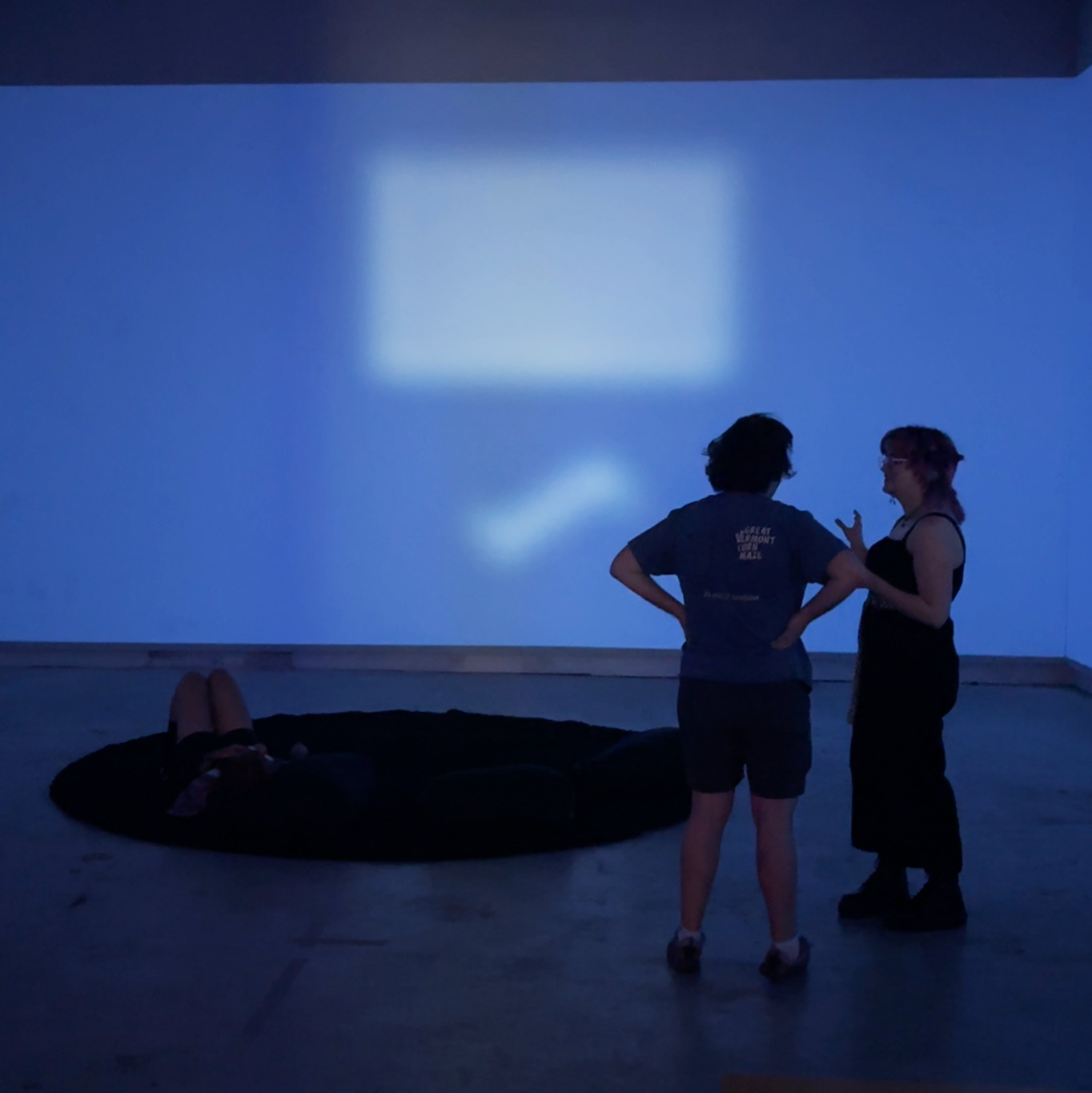 People standing and talking inside a blue-lit gallery space, while another person reclines on a dark floor installation beneath soft projected light.