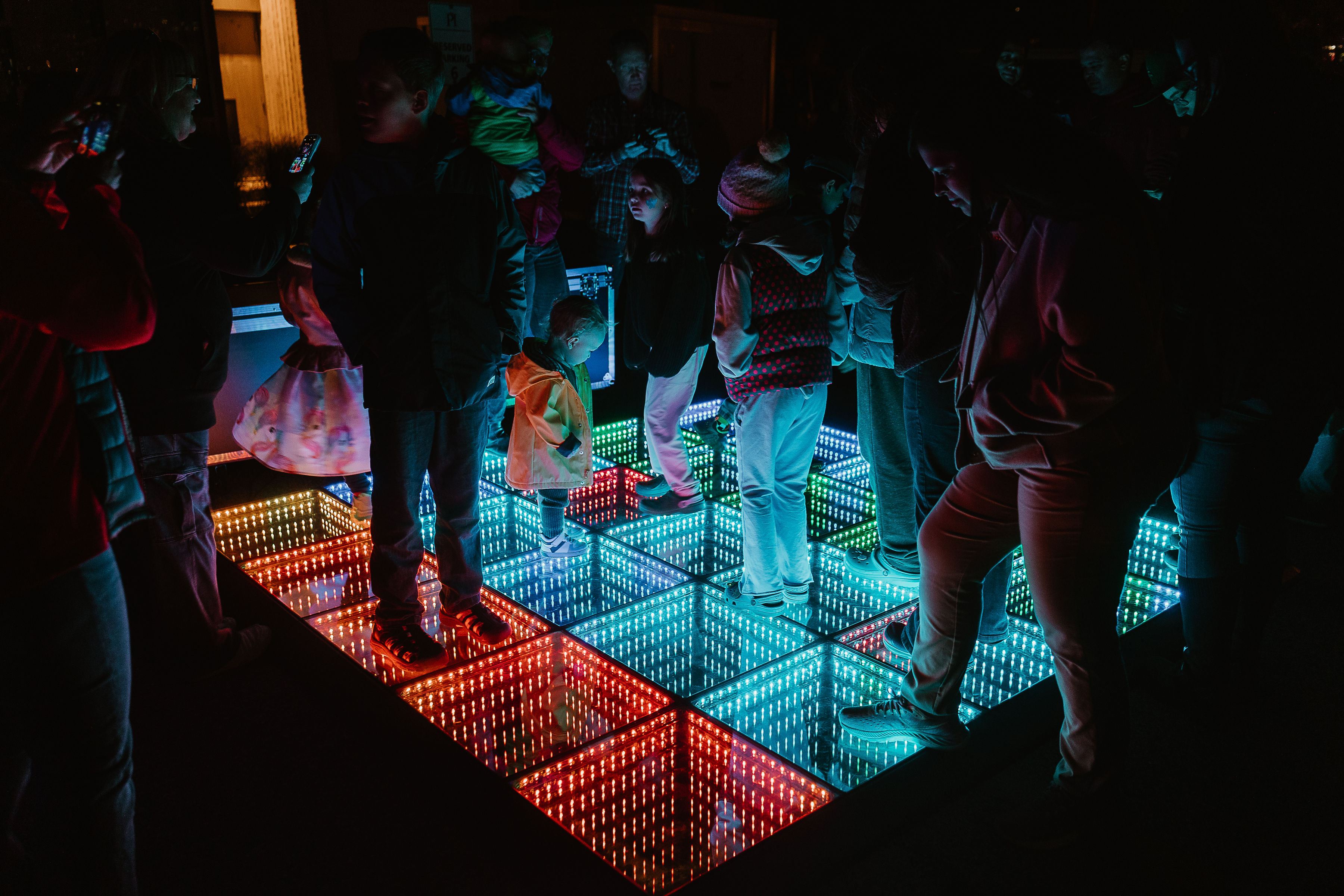 Festival visitors, including children, standing on an interactive illuminated floor made of glowing red, blue and green panels.