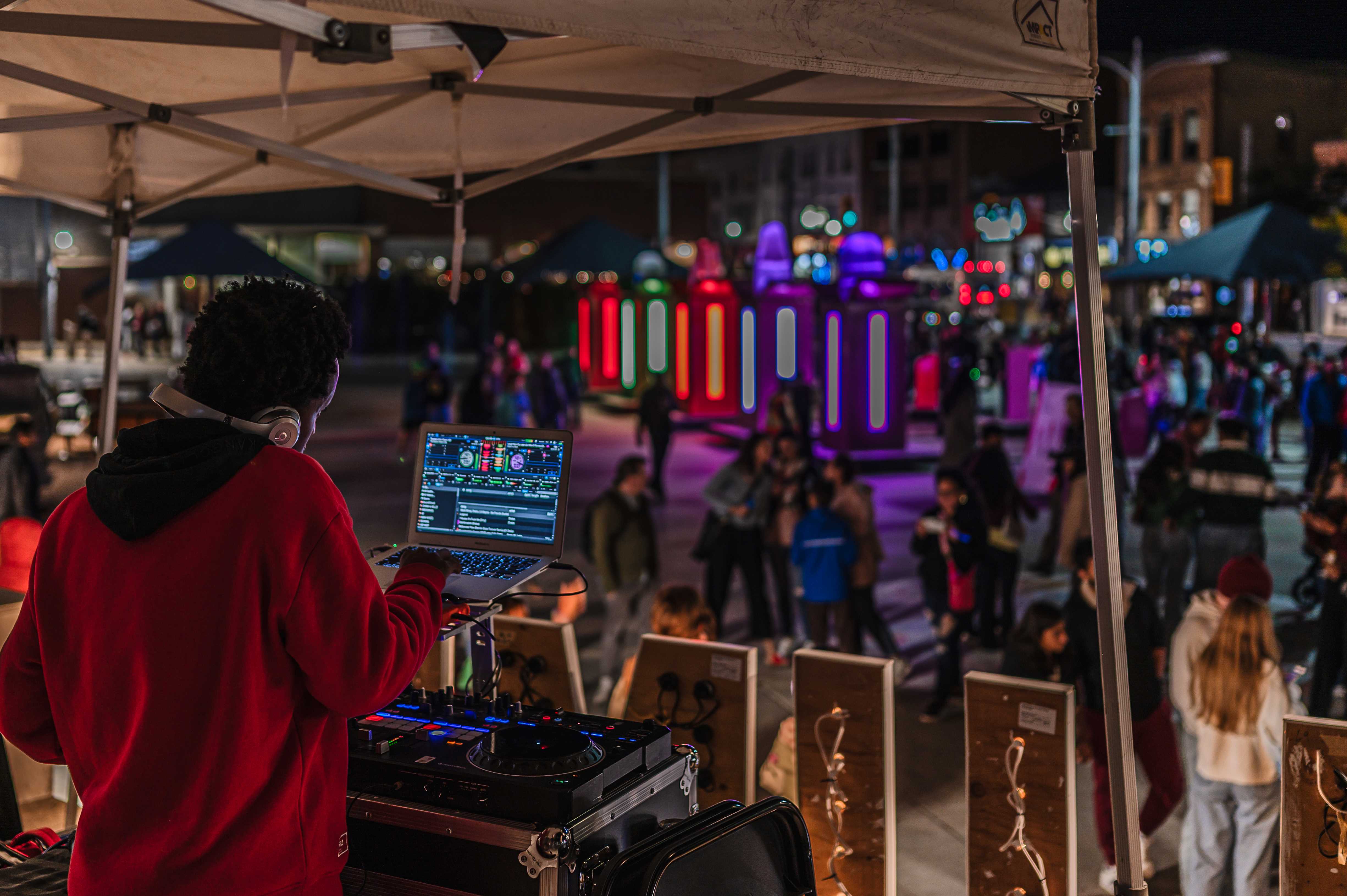 DJ performing under a tent overlooking a busy outdoor festival filled with colourful illuminated installations.
