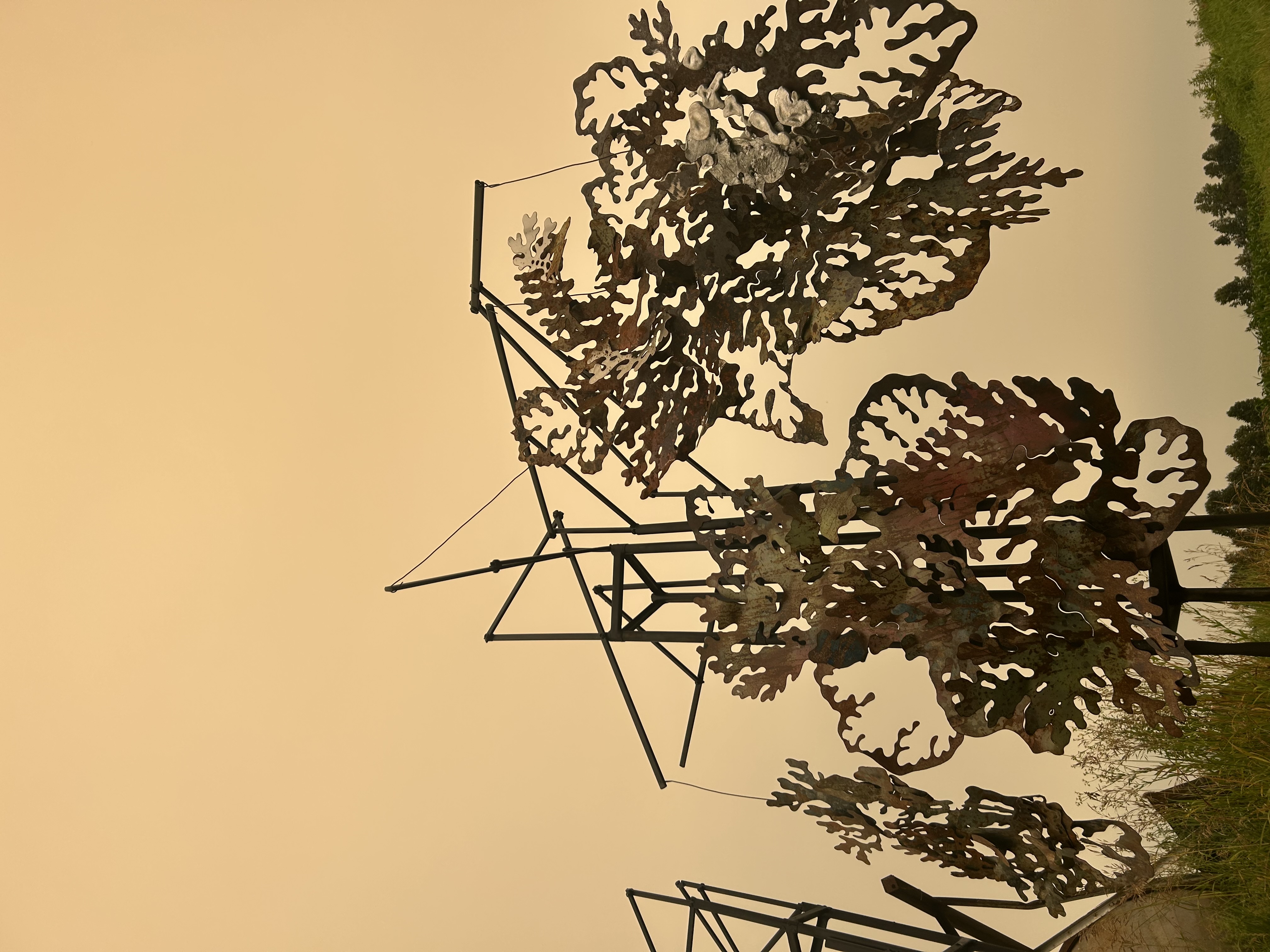Large outdoor sculptural installation made of intricate rust-coloured cut metal forms suspended from a black framework against a warm sky.