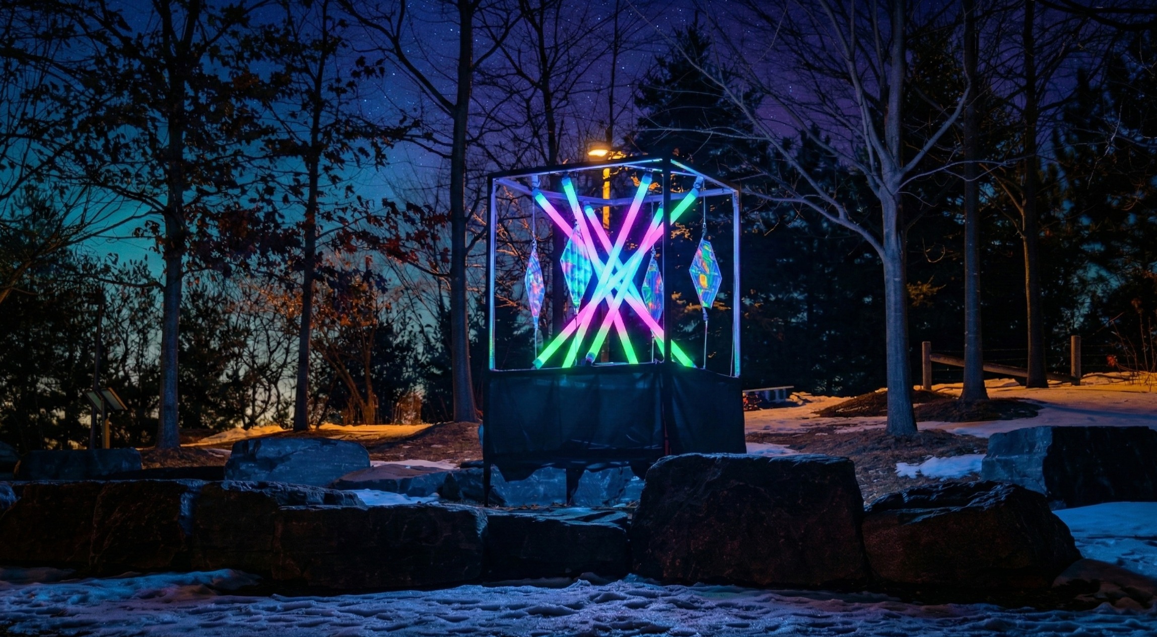 Colourful illuminated geometric sculpture glowing pink, blue and green among trees in a snowy park at night.