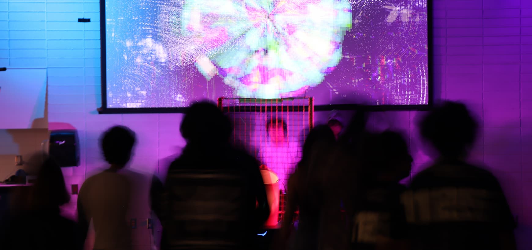 Audience members silhouetted in front of a large colourful projection during a live electronic music performance.