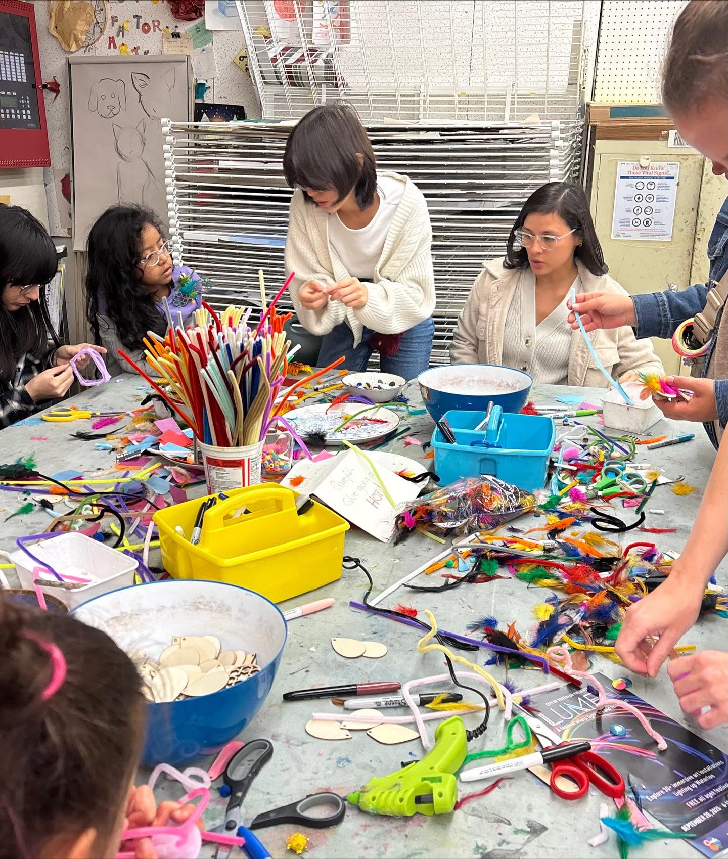 People gathered around a busy art-making table covered with colourful pipe cleaners, feathers, scissors, markers and other craft supplies.