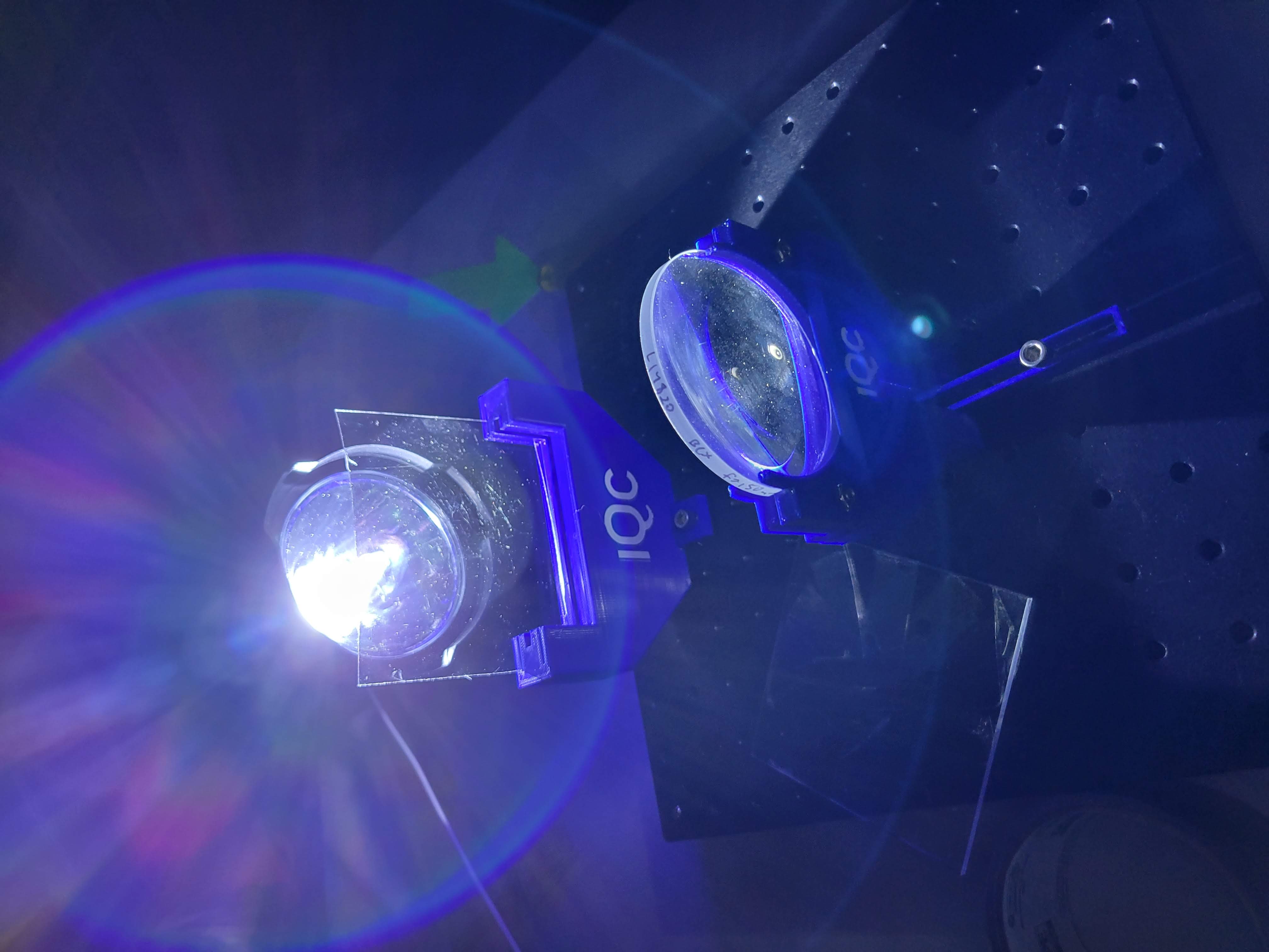 Bright white light shining through optical lenses and transparent panels, creating blue and purple lens flares in a dark space.