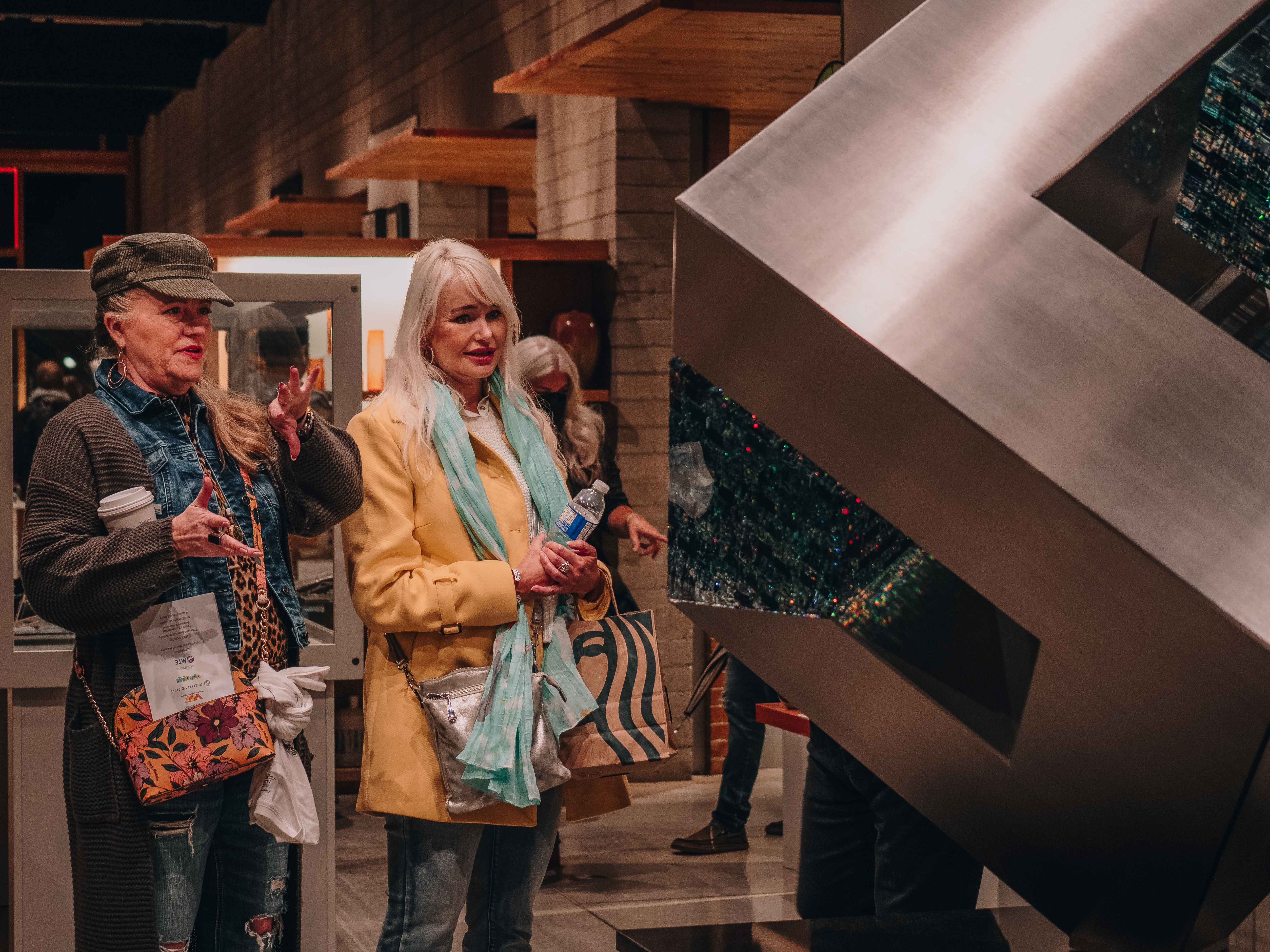 Two people view a large angular metallic artwork with dark reflective panels inside a gallery.