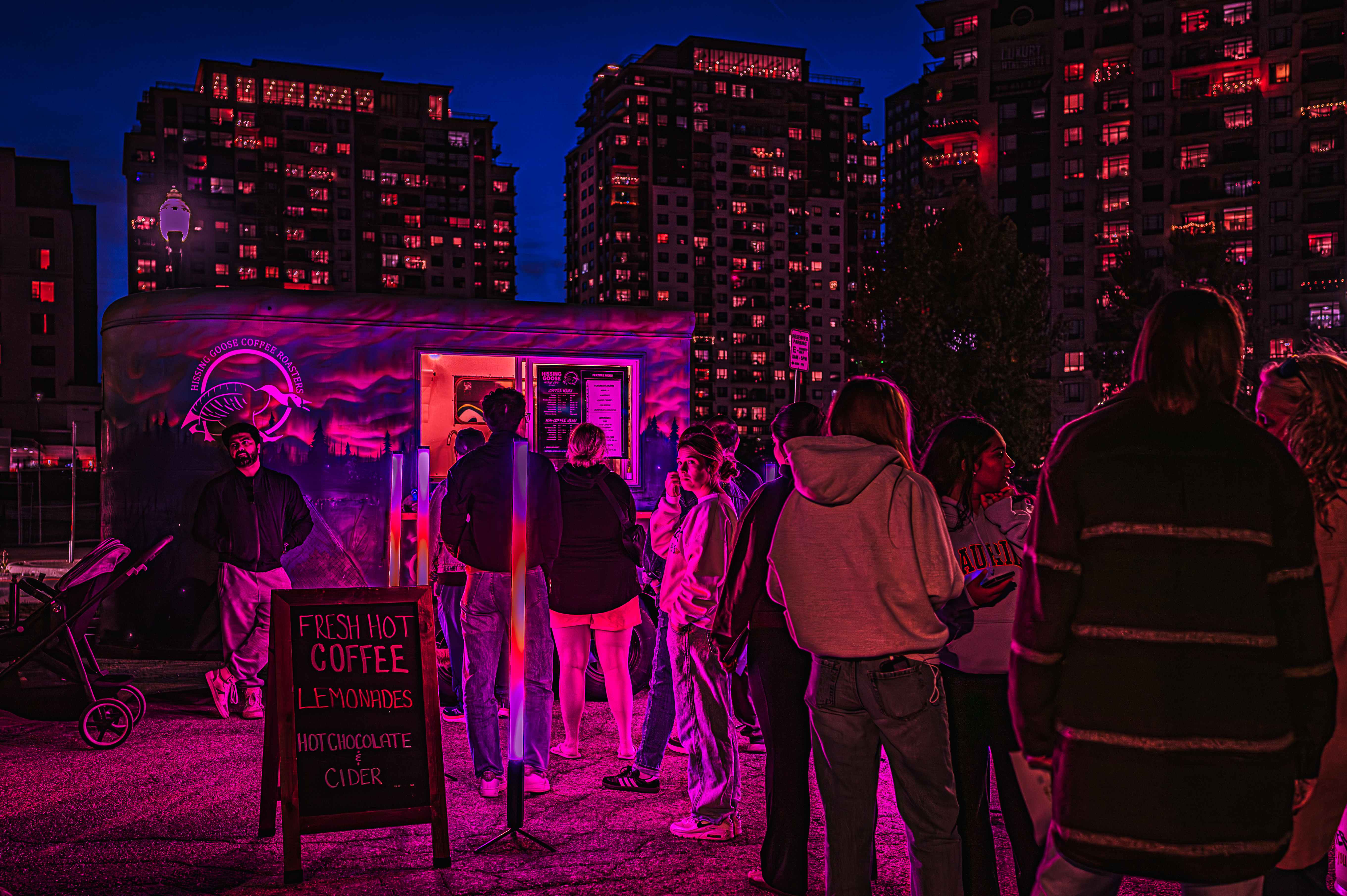 Festival visitors gathered around a brightly lit coffee trailer, with apartment buildings glowing pink in the background.