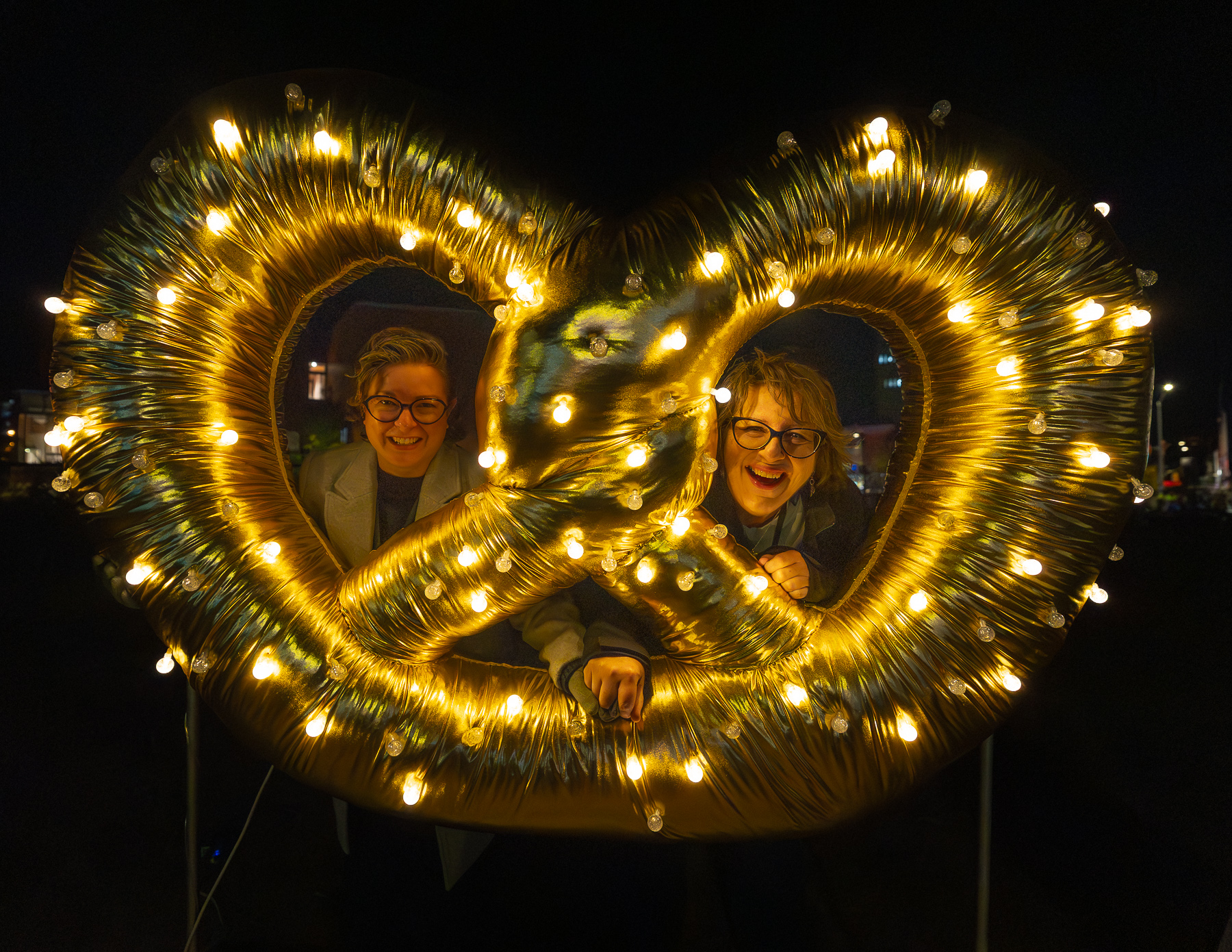 Two smiling people pose behind a large gold pretzel-shaped sculpture covered in warm white lights at night.