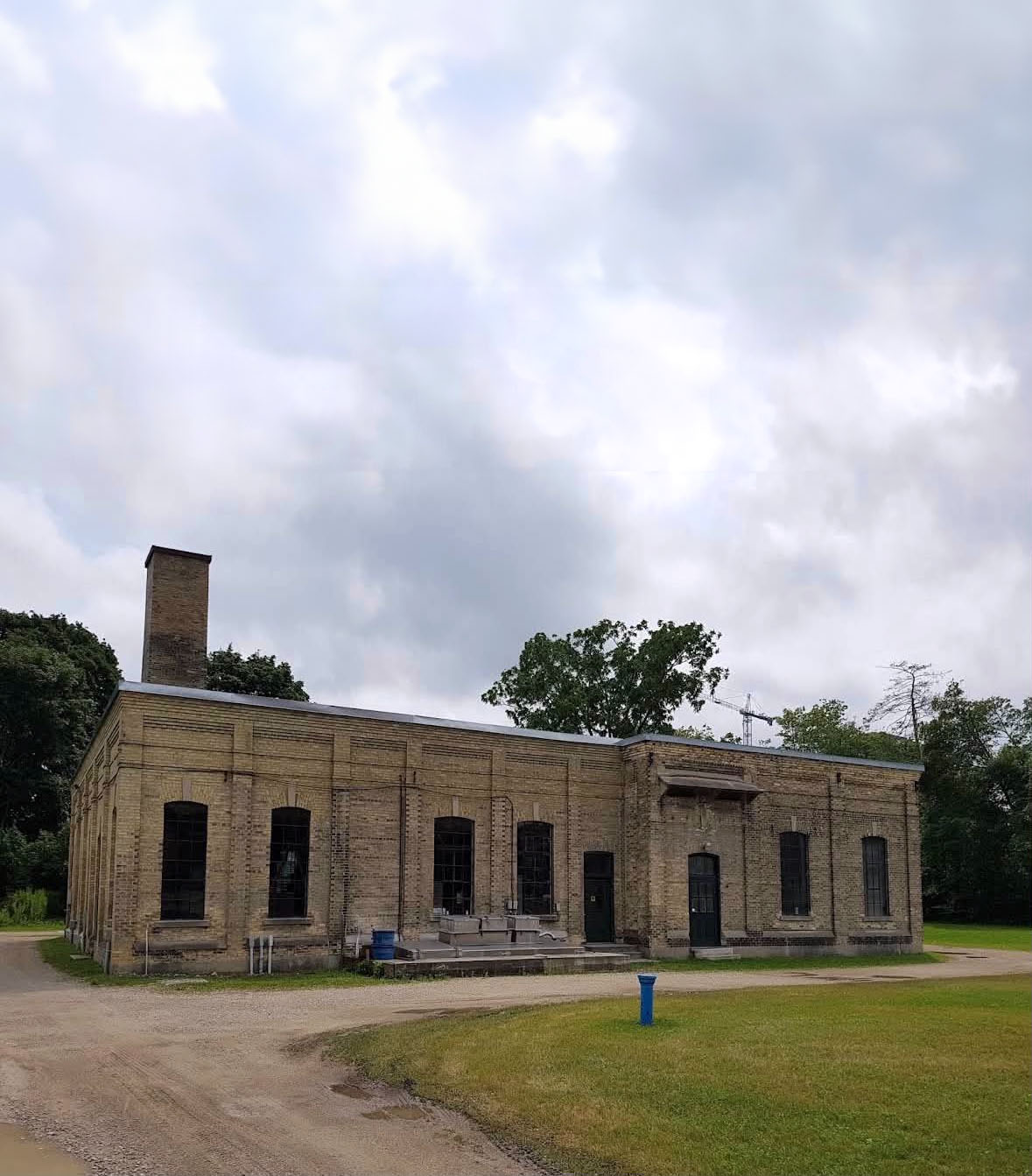 Historic William Street Pumping Station, a yellow-brick industrial building with tall arched windows and a large brick chimney, surrounded by grass and trees beneath a cloudy sky.