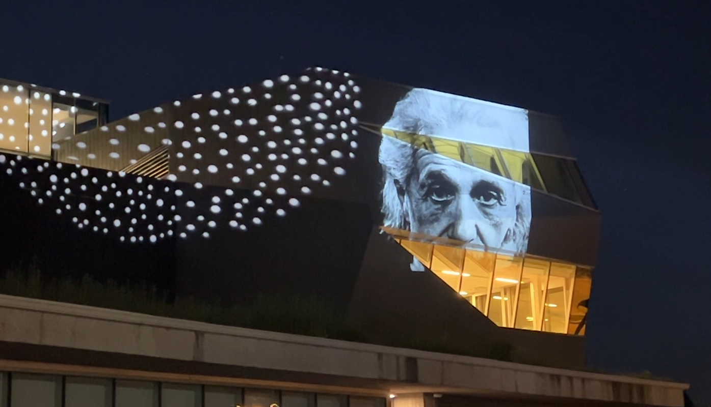 Nighttime projection artwork on a modern building, with clusters of white dots and a large black-and-white portrait projected across the façade.