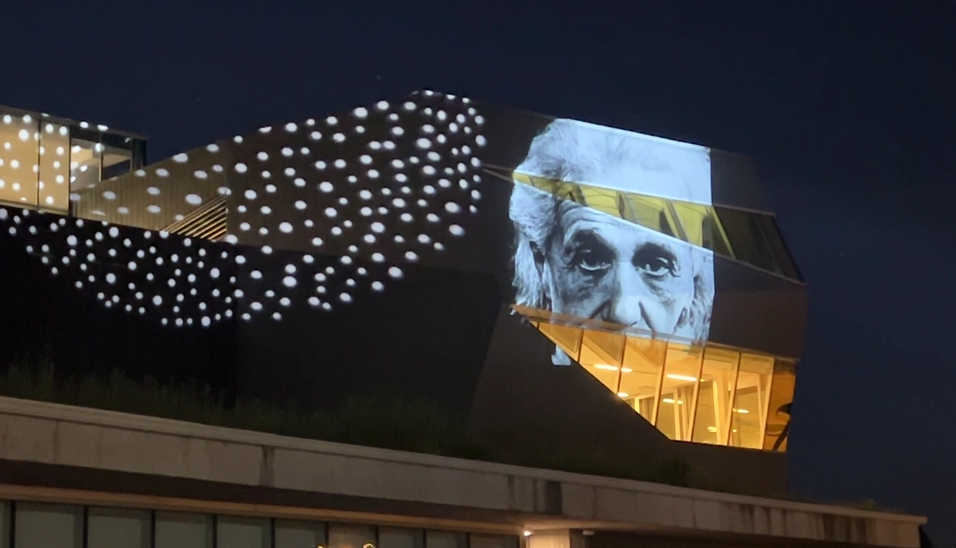 Nighttime projection artwork on a modern building, with clusters of white dots and a large black-and-white portrait projected across the façade.