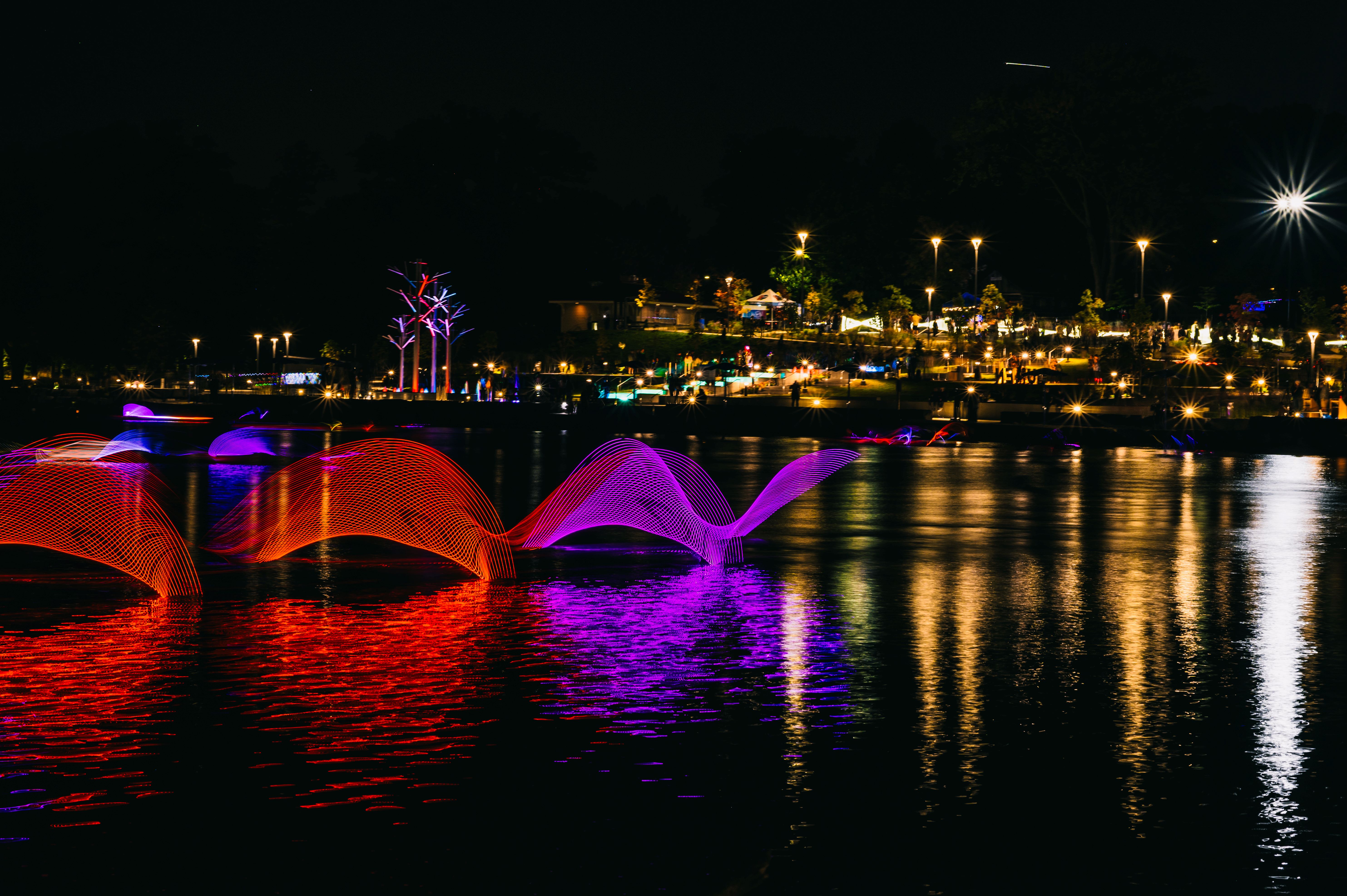 Night scene of a waterfront park with colorful light sculptures resembling waves in red and purple on the water, with illuminated trees and street lights reflecting in the water and people in the background.