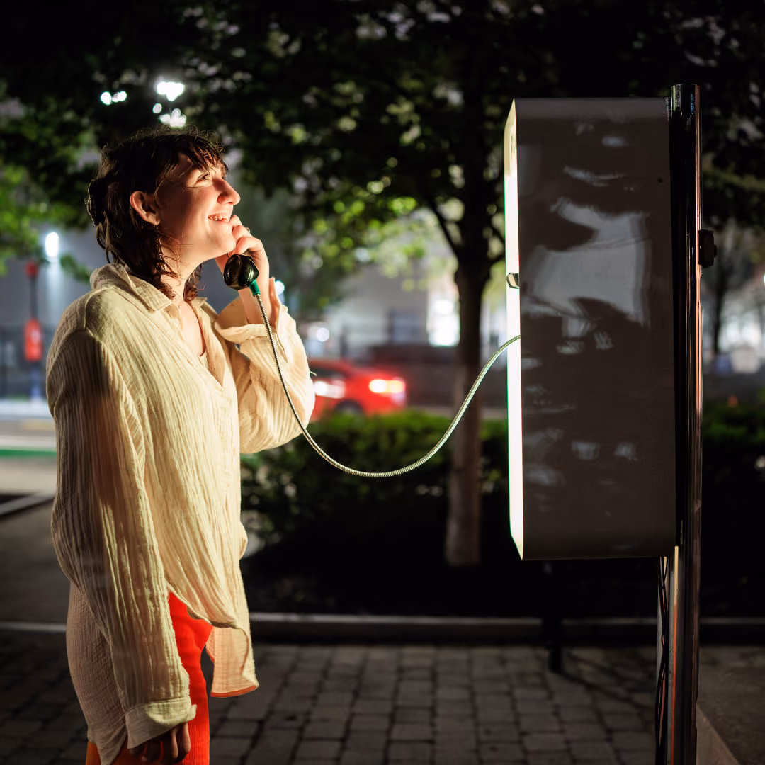 A person talking on a public telephone at night in an urban setting, smiling and looking upward.