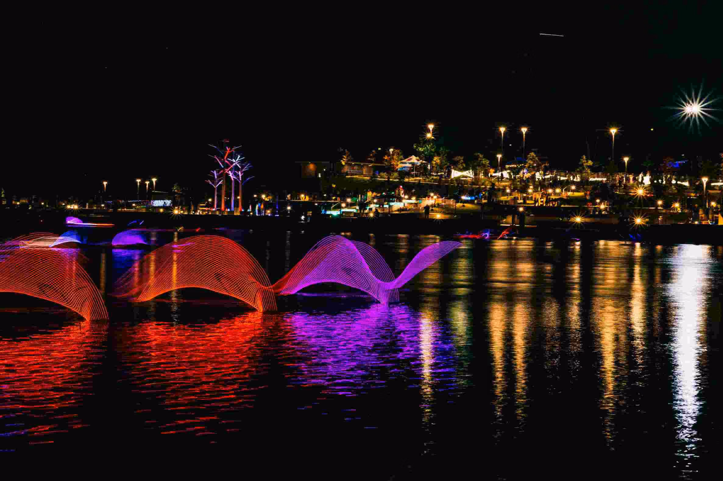 Night scene of a waterfront park with colorful light sculptures resembling waves in red and purple on the water, with illuminated trees and street lights reflecting in the water and people in the background.