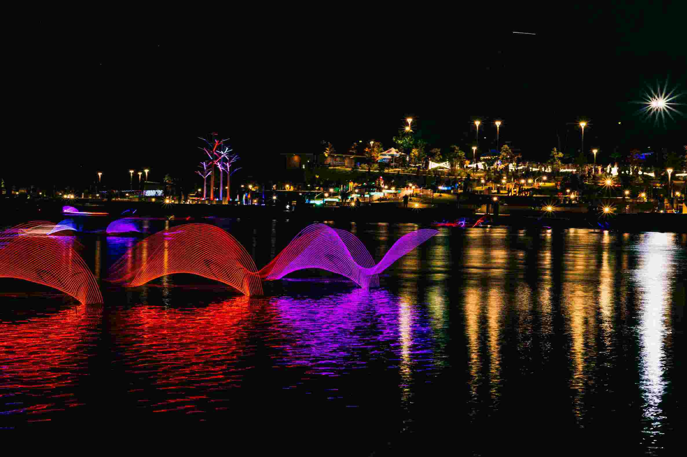 Night scene of a waterfront park with colorful light sculptures resembling waves in red and purple on the water, with illuminated trees and street lights reflecting in the water and people in the background.