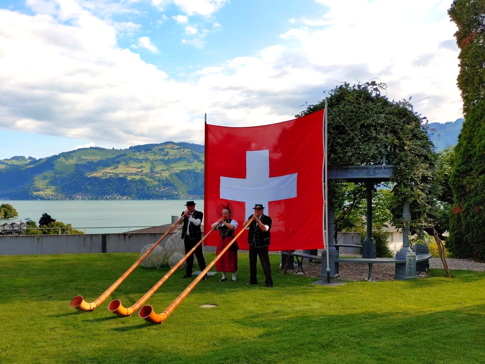 Alphornbläser vor Schweizer Flagge auf der Hotelwiese mit Blick über die Spiezer Bucht und den Thunersee.