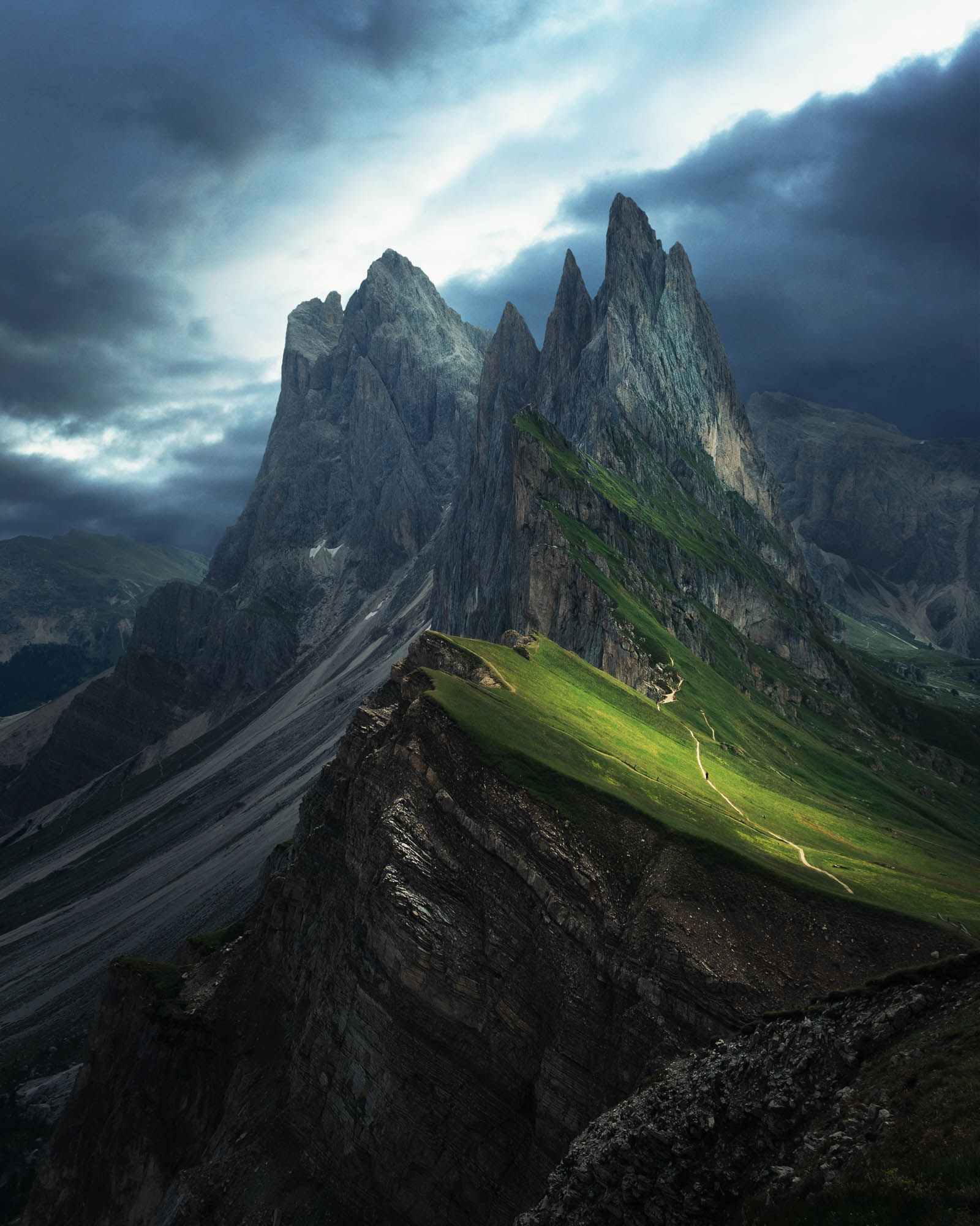 A picture of the Seceda mountain range in the northern Italian Dolomites. It is very dark and moody with light casting on the mountains. There is one man walking on the path.