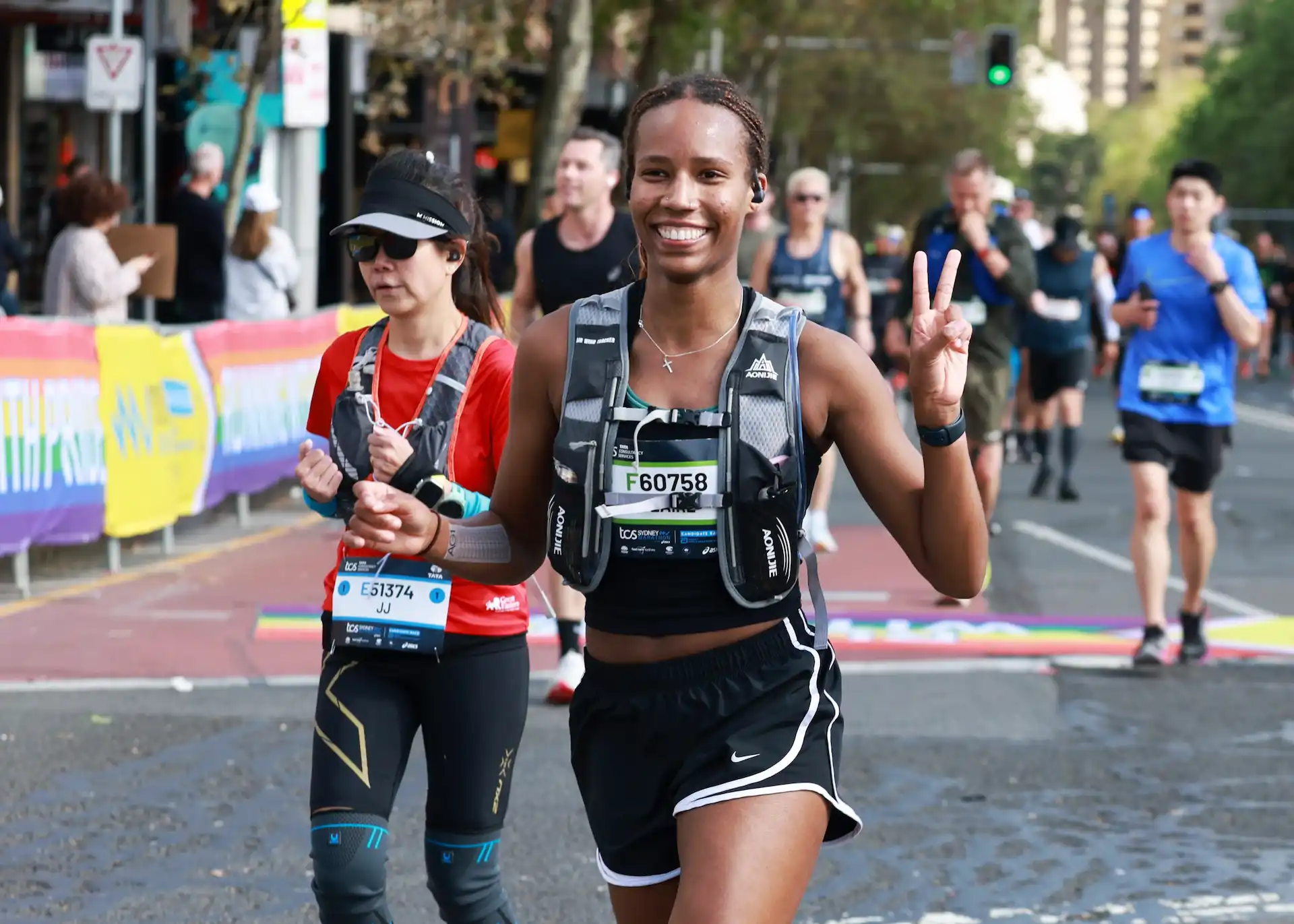 Two female marathon runners with race numbers, one making peace sign gesture on city street.