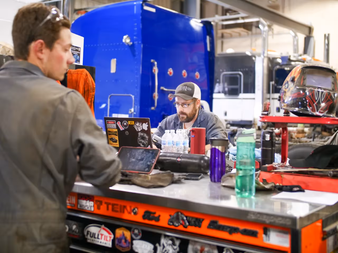Technicians at a workbench using laptops for heavy-duty truck diagnostics in a repair shop.