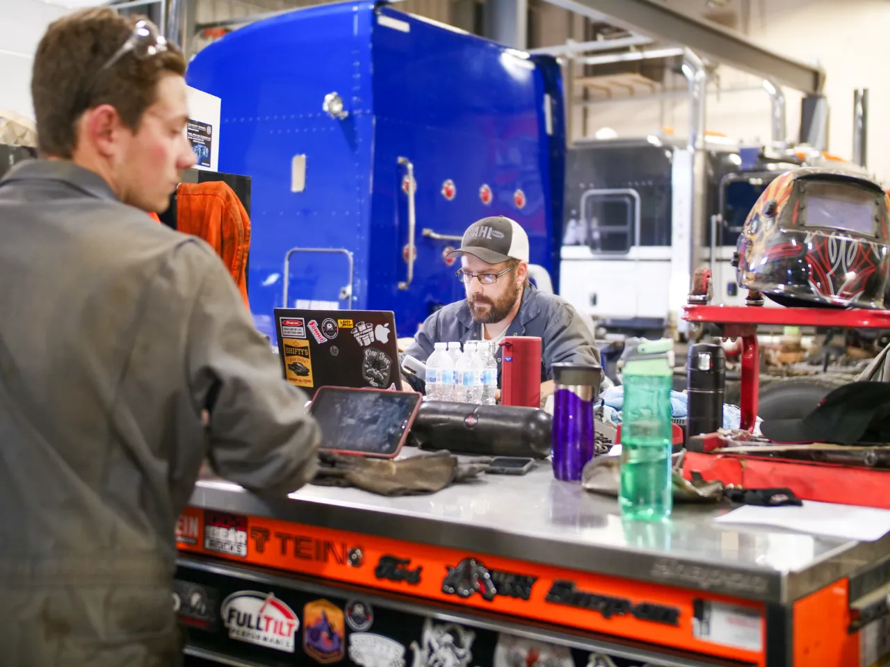 Technicians at a workbench using laptops for heavy-duty truck diagnostics in a repair shop.