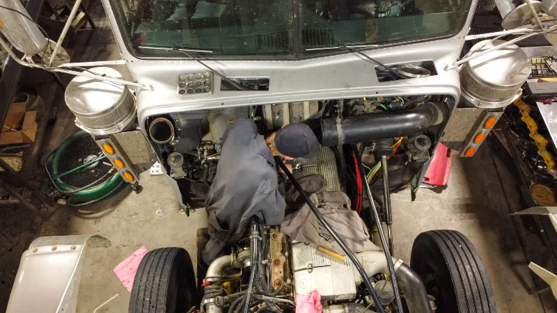 Technician performs engine repair on a heavy-duty truck with cab lifted and exposed diesel engine bay.