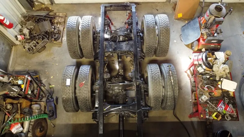 Chassis repair showing heavy-duty truck frame from above with dual rear tires, drivetrain exposed, and tools surrounding the work area.