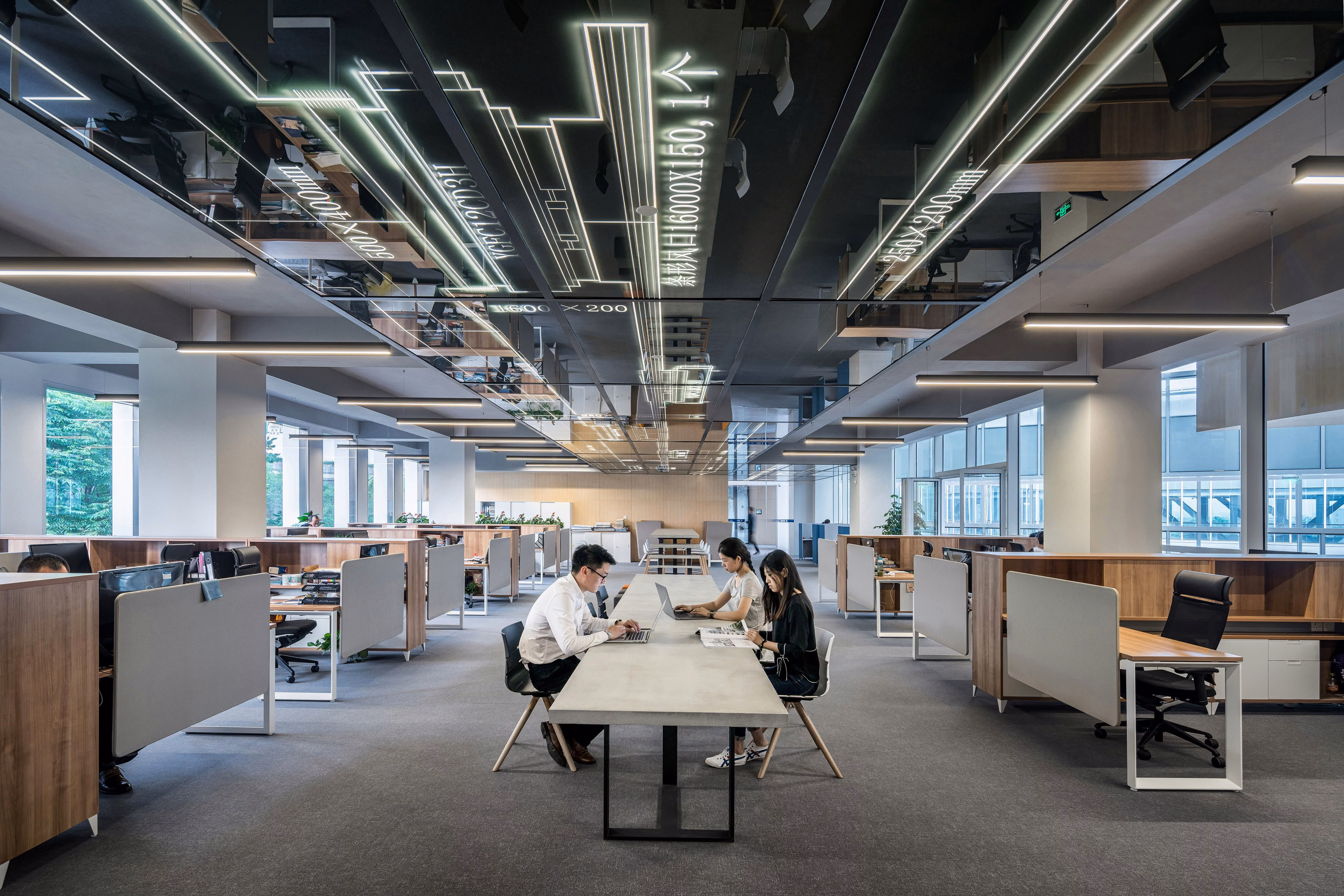 Modern office space with a long central table where three people work on laptops and documents; surrounding workstations are separated by gray partitions with wooden accents, under a ceiling with illuminated architectural design patterns.