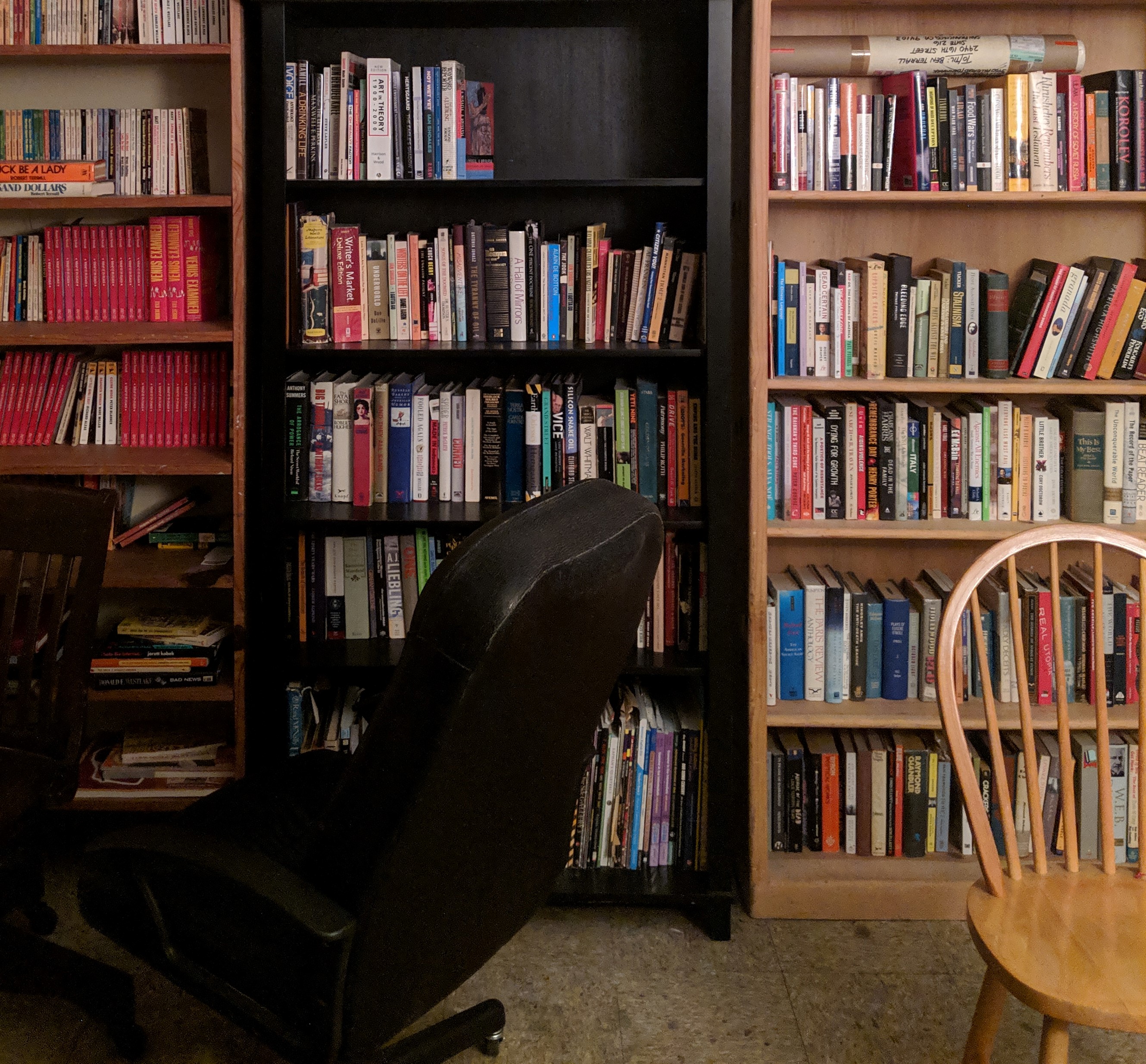 Three bookshelves filled with colorful books on the back wall of an art studio, with chairs in the foreground.