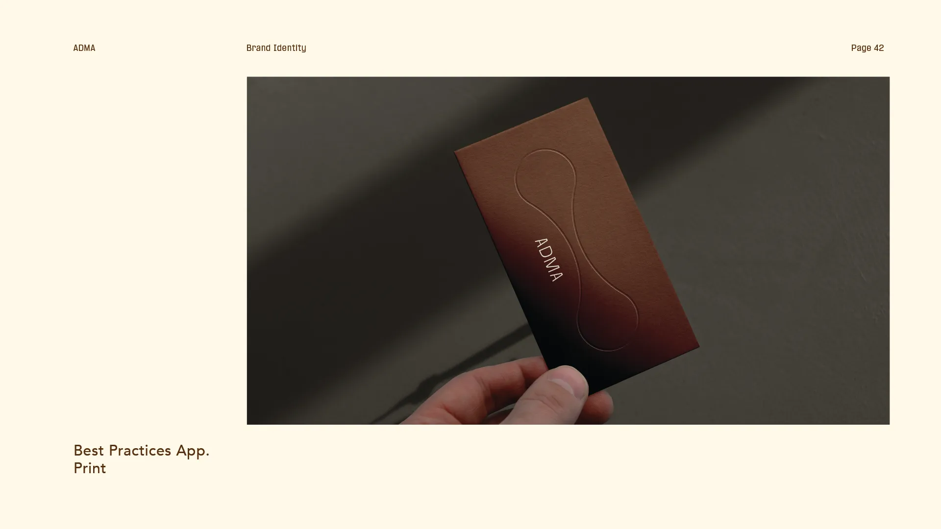 Hand holding a brown business card embossed with an abstract shape and the text ADMA, against a dark background.