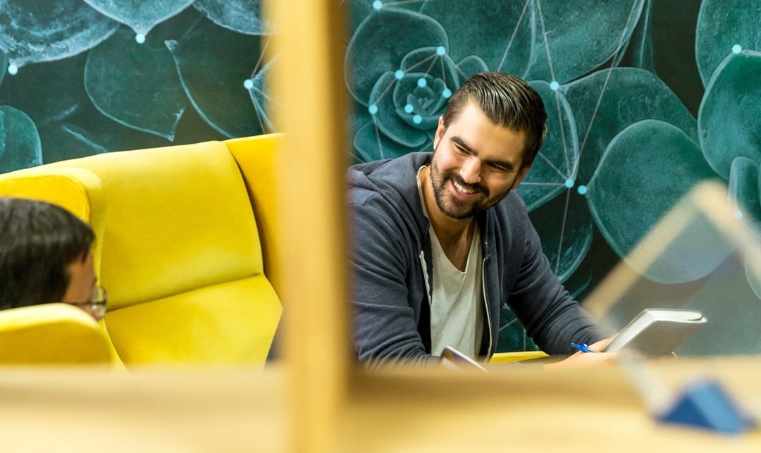man smiling while sitting on a couch holding what seems an agenda. He is looking to a friend or colleague that is sitting on another couch beside him