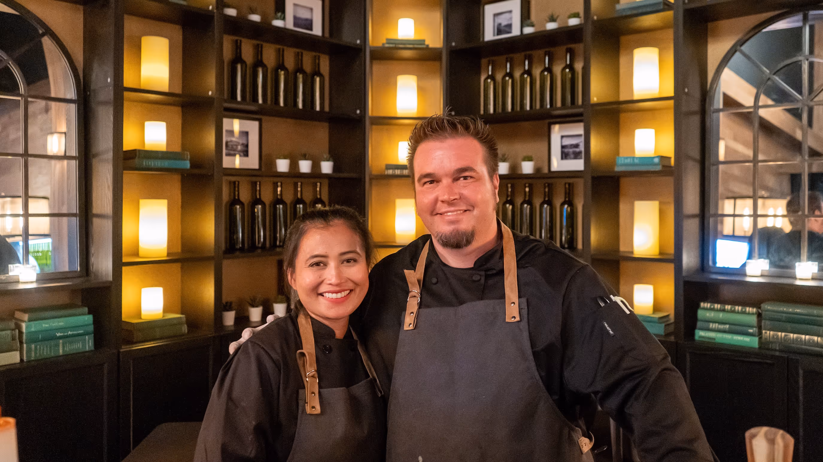 Two chefs in black uniforms and aprons smiling in front of illuminated shelves with bottles, books, and candles.