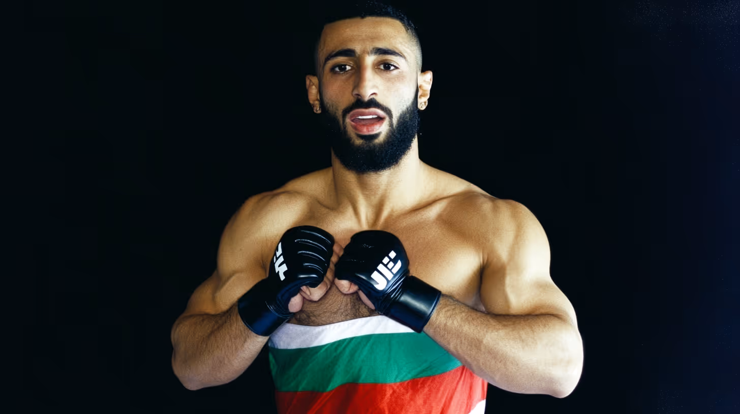 MMA fighter with crossed arms and black gloves, bare muscular torso, and a green-red striped outfit against a dark backdrop.